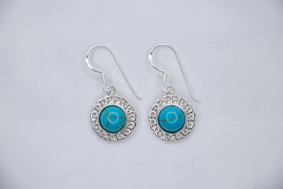 A Turquoise Serenity Drop Earrings featuring a vibrant turquoise gemstone set in a delicate sterling silver frame with an ornate design, top views.