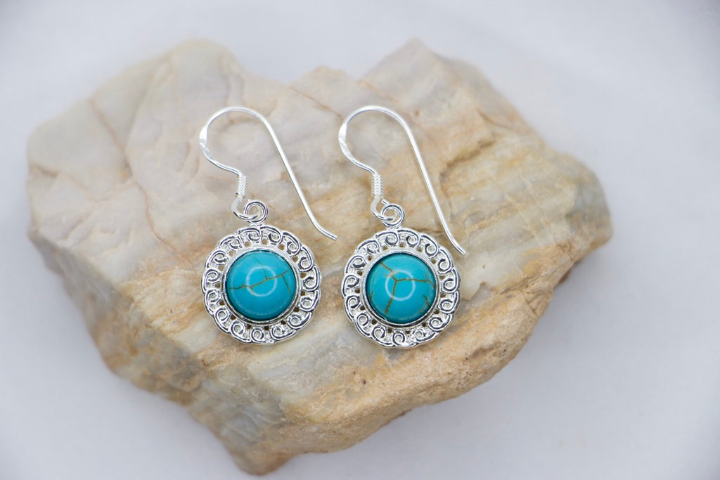 A Turquoise Serenity Drop Earrings featuring a vibrant turquoise gemstone set in a delicate sterling silver frame with an ornate design, rock views.